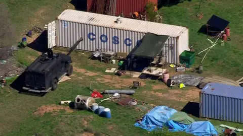 Australian Broadcasting Corporation A container on a grassy area surrounded by empty barrels, tarps and a black police vehicle