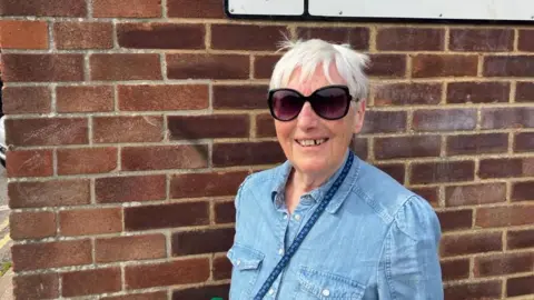 Vikki Irwin/BBC Margaret Hancock is standing by a brick wall and is smiling at the camera. She is wearing dark sun glasses and a blue denim shirt. 