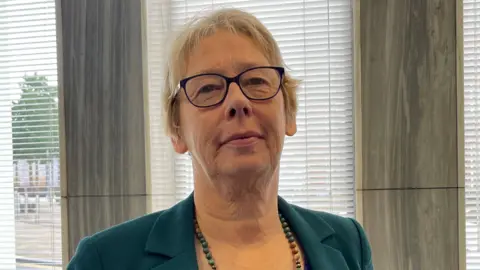 Jane Ashworth standing in an office with venetian blinds on the windows. She has short fair hair and is wearing glasses, a dark green jacket and beaded necklace.