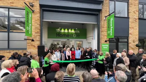 A crowd of people outside the Thames Hospice Homestore for it's grand opening with Anita Dobson. A green ribbon is about to be cut by Anita Dobson dressing in a red jacket and scarf.