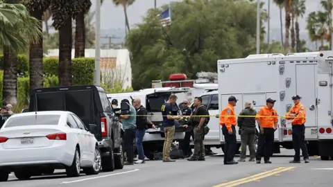 First responders in FBI shirts and orange uniforms stand on a palm tree-lined street. There are many trucks and a section of the stree is blocked off with yellow caution tape.