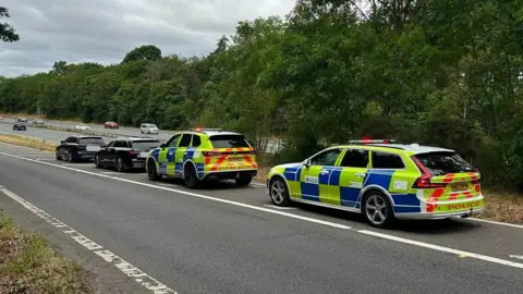 Police vehicles with flashing lights are parked on the roadside of the A46 near a wooded area while officers carry out a traffic enforcement operation, with several stopped cars lined up ahead under an overcast sky.