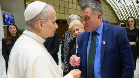 Ian Byrne, wearing a tie, blue shirt and dark blue suits, meets the Pope, dressed in all white. there are other people stood in the background.