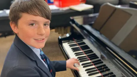 River sat at a piano wearing a navy jacket and blue shirt with his blue and red school tie. He has short brown hair and brown eyes.