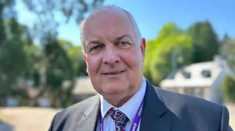 Raymond Bremner is wearing a grey suit with a red patterned tie and a purple lanyard. He is standing in front of a blurred-out background that includes houses and trees. 