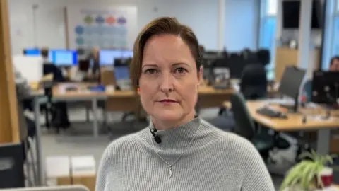Alison Hernandez with a stern expression wearing a grey jumper, standing in an office