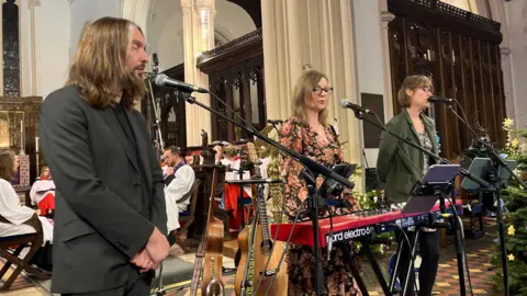 Jon Wright/BBC A man and two women sing into microphones at the front of a church. The woman in the middle has a keyboard in front of her, and there are guitars on stands next to her.