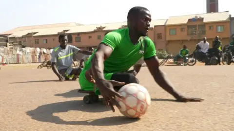 A skate soccer player on a board wearing green goes to play the ball with his right hand with another player just behind him as a small group of spectators resting on motorbikes watch the action on a hard-looking, dry African court