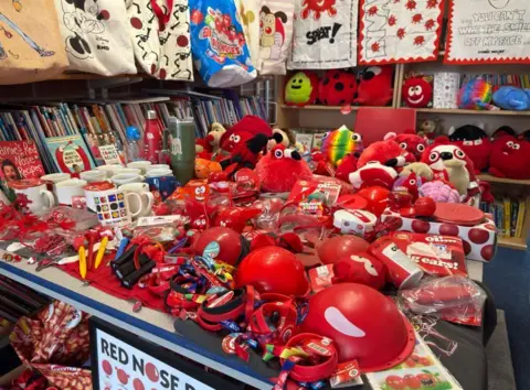 Rob Rose/BBC A vast array of Comic Relief items on a table include red noses, pens, wristbands, soft toys, tea towels, tote bags, cups, mugs, erasers, water bottles and keyrings.