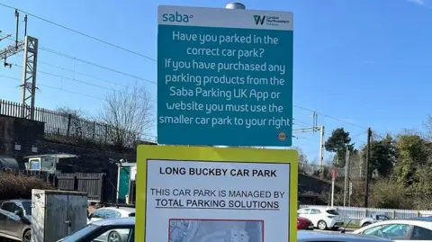 Total Parking Solutions Sign in a car park in white writing with blue background, which states that drivers who have paid Saba must park in the smaller car park. It includes a diagram.  There are cars parked behind the sign and a railway line is visible to the left.