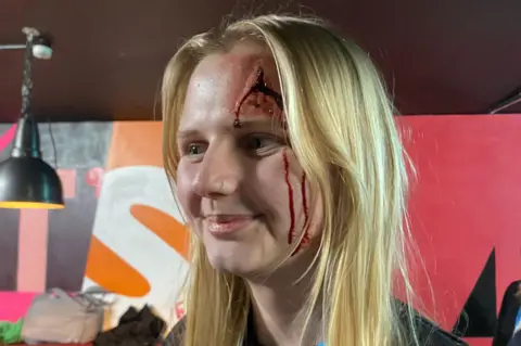 BU A young woman with long blonde hair has special effects make up on her face, giving the appearance of a bloody gash in her forehead.