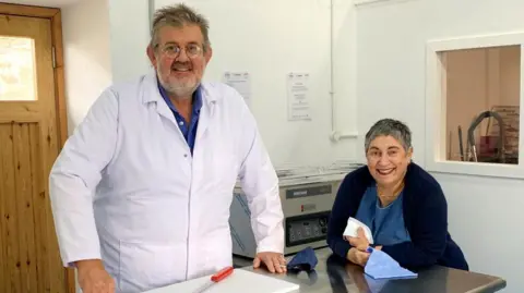 Richard Irvine Richard Irvine stands behind a metal counter with a white chopping board and red-handled knife. He has grey hair and a beard and is wearing spectacles, a royal blue shirt and a white overcoat. His wife leans on the counter next to him, wearing a navy cardigan, a blue dress and dark red lipstick. Her hair is grey and cropped short.