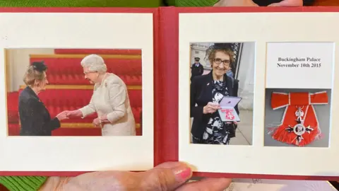 Tom MacDougall/BBC A small red book of photos, showing Meg meeting the Queen and photos of the medal she was awarded. They are dated November 10th, 2015.