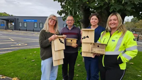 Debbie Morrell and Kev Klein from Friends of Alderman Kneeshaw Park, Jill Pawson, charge nurse for the Day Surgery Centre, and Amy Lockyer, Commissioning and Support Services Manager for NHS Humber Health Partnership. All smiling and holding different sized wooden homes, some with bat drawings on, for wildlife in front of the day surgery centre.