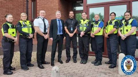 Nine people stand next to each other in a line smiling at the camera. Most of them have police uniforms on, one of them that of a senior officer, and one man is wearing a suit.