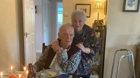 Eiry Miles A man with grey hair around the side of his head, a checked shirt and purple cardigan sat at a table with a plate in front of him and some candles. There is a walking frame in the right of the shot. Behind him is a woman with short grey hair with hands on both of his shoulders