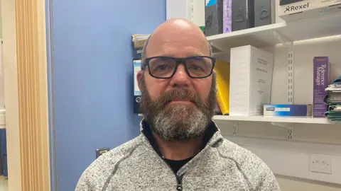 Neville pictured looking into the camera. He's wearing a grey quarter-zip fleece and a pair of black-framed glasses. He's pictured indoors inside an office, which has blue walls, and to his left are a set of white shelves which have black, yellow, and white files and folders on them. There's a set of yellow curtains hanging on the left of the image.