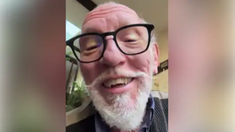 A close-up selfie of an elderly man with a white beard and glasses. He is sitting in a car.