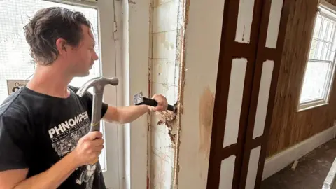 Francesca Griffiths A man in a black T-shirt holds a hammer in his right hand, a chisel in his left and has the chisel against a wall as he prepares to hit it with the hammer. The wall is at the side of a doorway inside a building.