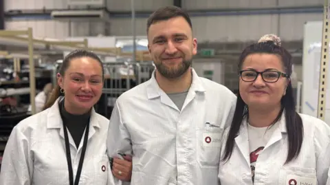 Three people stand side by side on a factory floor - they're all wearing white lab coats. Michael Smola is the tallest and is in the middle of the three. He has short brown hair and a cropped beard. He's smiling at the camera. To his left, his partner Natasha is linking arms with him. Her hair is tied back in a low pony tail and she is smiling. Michal's mum Monika is standing on his other side. Her hair is also tied back and she is wearing dark rimmed glasses and smiling. 