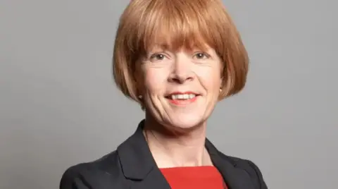 Parliament MP Wendy Morton in her official parliament photo. She is against a plain, dark background, wearing a blazer and a smart red top. 