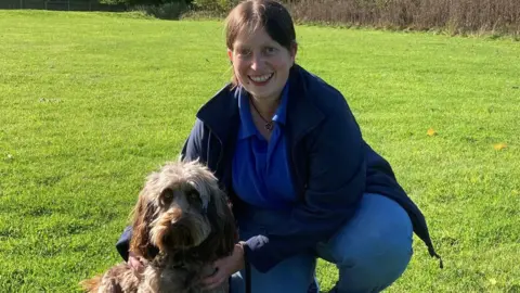 A woman who is crouched down on a big patch of grass holding a brown cockapoo. The woman is wearing light blue jeans, a blue polo shirt and a navy fleece. She is smiling into the camera. The dog is also looking straight into the camera and the sun is shining onto the grass.