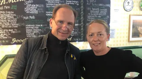 Carly Lea Ralph Fiennes, wearing a dark denim jacket over a black zip up hoody and glasses, stands next to Carly Lea, who has blonde hair tied back and a black long-sleeved top. 
