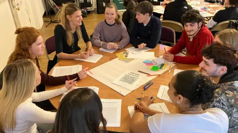 Jon Wright/BBC Ten sixth form students sit around a table with pens and paper in the middle.