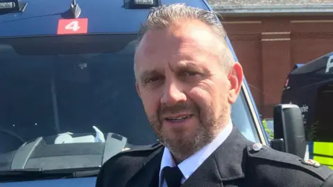 Guernsey Police Damian Kitchen stood in front of a blue vehicle. He is wearing a black jacket with two buttons on each shoulder. He has short hair, a beard and is smiling