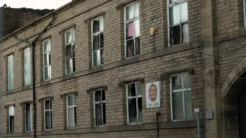 Mark Graham/BBC A two-storey stone-built commercial building with multiple broken and damaged windows is shown from street level. Numerous window panes on both floors are shattered or missing entirely, with some openings partially covered by curtains, boards, or remaining fragments of glass. Electrical cables are visible running across the exterior wall near the upper floor. A weathered sign mounted on the front of the building reads "new weighing line" with a telephone number beneath it, although parts of the text are difficult to read. The building appears vacant or neglected, and the surrounding stonework shows signs of age and wear. An arched stone entrance is partially visible on the right-hand side of the image.