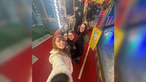 Gemma Phillips Five women taking a selfie, smiling to camera, in an amusement arcade