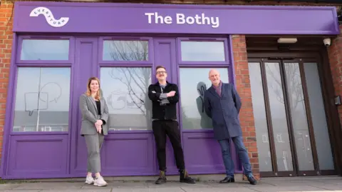Everyturn Three people, Mayor of the North East Kim McGuinness, Everyturn CEO Adam Crampsie and James Duncan from CNTW NHS Foundation Trust, stand in front of a purple building with a sign reading The Bothy.