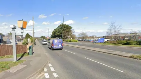 The back of a speed camera can be seen on the left aimed towards a dual carriageway that has a van and car driving on it. A retail park can be seen in the background. 