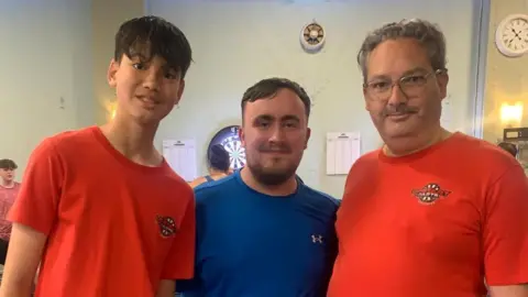 Upstart Darts Three men stand together in a hall. The teenager to the left has dark short hair and wears a red t-shirt with a darts logo on the left breast. The man on the right wear a matching t-shirt and wears glasses, with a moustache and grey curly hair. The man stood between them both is famous darts player, Luke Littler, wearing a blue t-shirt with a soft brown beard and short hair.