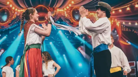 A group of young people folk dancing on the stage at the Eisteddfod in Pontypridd 2024. They are all in traditional outfits. There is a man and woman on the front of the stage with their hands in the air clapping whilst smiling at each other.