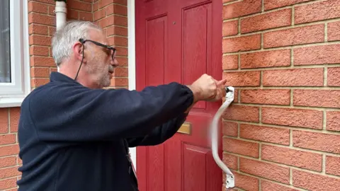 Tom Jackson/BBC Roger Hewish fitting a white grabrail to the side of a red front door on a brick house. He has white hair and a white beard, wearing glasses and a blue fleece.