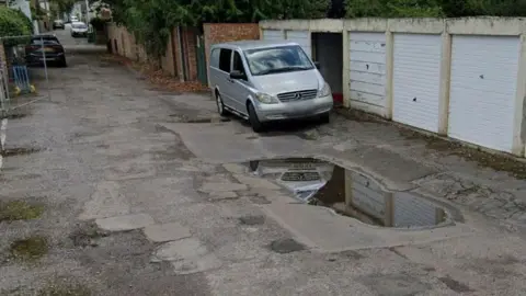 Google A bumpy, patched up back road where there is a row of garages with white doors and a silver Mercedes van. There is a large puddle due to the poor road surface.