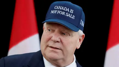 A close-up image of Doug Ford, who is looking to the left and is wearing a navy cap that reads "CANADA IS NOT FOR SALE" in white letters.