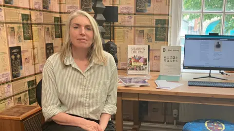 A woman in a green and white striped shirt sits in a room with lots of posters. There is also a computer on a desk. 