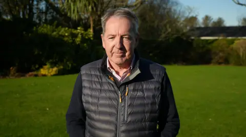 Tim Crowley - A man standing in a garden wearing a black gilet and navy cardigan underneath.