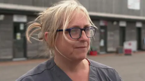 A woman with blonde hair that is tied up with loose strands around her face is pictured by a row of buildings. She has glasses on and is wearing a grey coloured top.
