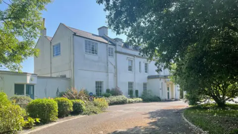 A white manor house with a curved driveway and trees and plants outside.