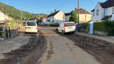 Two vans parked either side of the road on Heol Berry, Gwaelod y Garth. There is mud on either side of the of the road on the residential street.