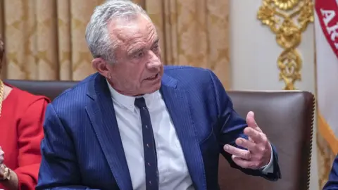 EPA/Shutterstock Robert F. Kennedy Jr siedzi przy stole i macha ręką do osoby siedzącej obok niego. Ma na sobie niebieski garnitur w prążki i wąski krawat. W tle widać złoto, a po prawej stronie ramię osoby w czerwonej sukience.
