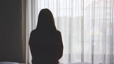 A woman with long dark hair sits in the dark on the edge of a bed facing a window with white net curtains drawn with her back to the camera