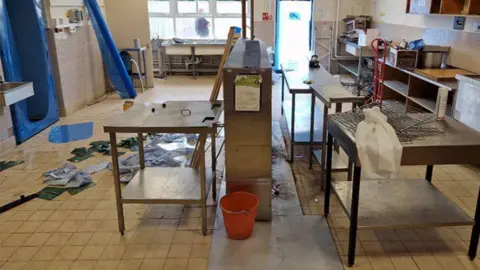 The inside of the school kitchen during construction. There are metal tables and grills in the centre of the tiled floor. The room is messy and there are paper towels all over the ground.