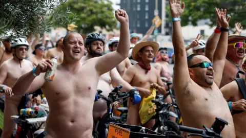This image as a hundred of half naked people waving their hands while sat on a bicycle. Most of the identifiable people in this picture are men. One man is holding a drink bottle and most of the others are wearing helmets with sun glasses. 