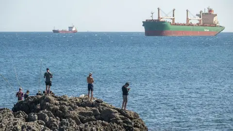 A group of men are seen from the back on a rocky outcrop with the blue sea and large ships in the background, on 22 August, 2026 in Ceuta, Spain.