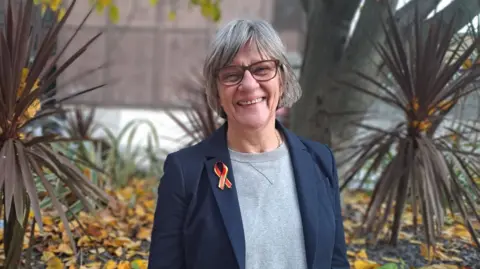 Samantha May, helpline manager, looking at the camera and smiling in a garden with plants and trees in it. She is wearing a blue blazer and a grey top underneath.