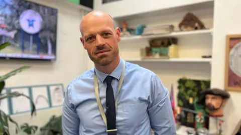 A man in his 30's with a bald head and slight facial hair wearing a blue shirt with a navy blue tie